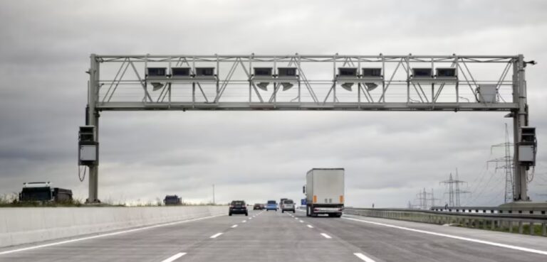 Cars and a truck driving on a highway under a gantry with cameras and sensors, cloudy sky above. tolls